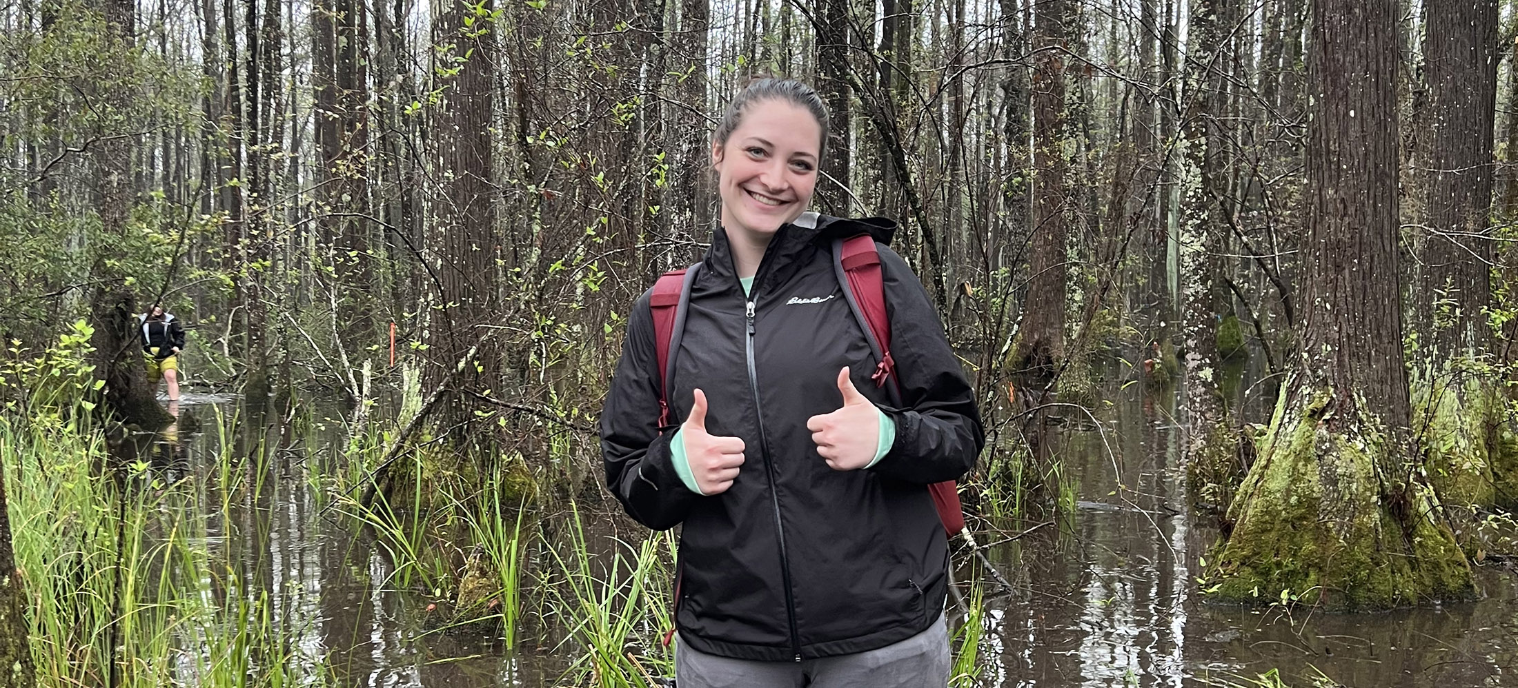 A young woman stands in a swamp surrounded by trees