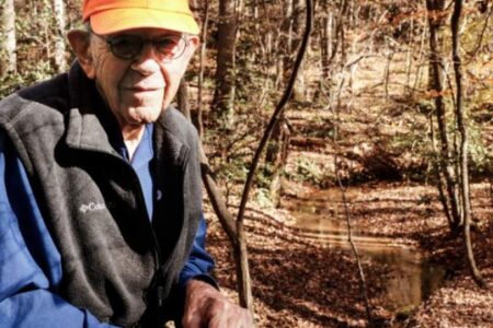An elderly man wearing an orange cap and dark vest stands outdoors in a sunlit forested area near a shallow creek.