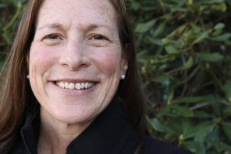 A woman with long brown hair and a black jacket smiles outdoors in front of green leafy bushes.