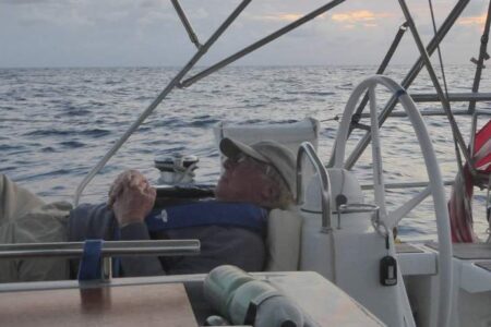 An older man wearing a cap and life vest lies on a sailboat deck at sea, relaxing with hands folded, while the sun sets in the background.