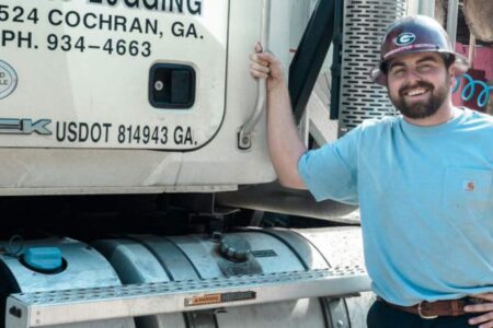A man in a blue shirt and hard hat stands next to a logging truck labeled with company information and phone number in Cochran, Georgia.