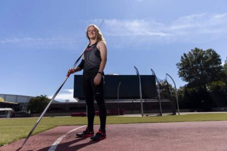 A person stands on a track holding a javelin, dressed in athletic wear, with a field and stadium equipment visible in the background under a clear sky.