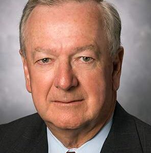 An older man with gray hair in a suit and red tie poses for a formal portrait against a gray background.