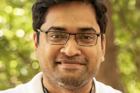 A man with glasses and a short beard, wearing a white shirt, smiles at the camera outdoors with greenery in the background.