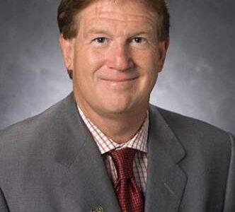A man in a gray suit, checked shirt, and red tie poses for a professional portrait against a gray background.