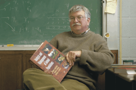 A man with gray hair and glasses sits in a classroom, holding a textbook titled Analysis and Management of Animal Populations, with a chalkboard in the background.