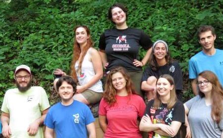 A group of ten people pose outdoors in front of green foliage, with some standing and some seated or kneeling on a low wall.
