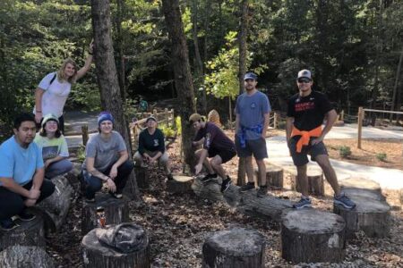 A group of people pose outdoors on large tree stumps in a wooded area with a gravel path and trees in the background.
