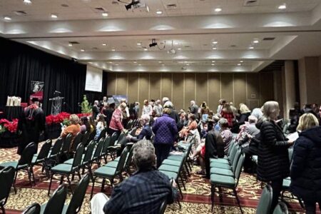 A group of people gathers and chats in a large conference room with rows of chairs, a stage, and holiday decorations.