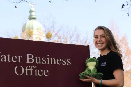 A woman stands next to a Water Business Office sign, smiling and holding a green stuffed frog. A dome-shaped building is visible in the background.