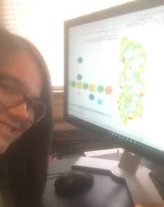 Person with long hair and glasses sitting at a desk in front of a computer displaying a colorful data chart or diagram.