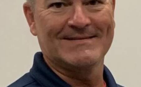 A middle-aged man with short hair is wearing a navy collared shirt and smiling at the camera against a plain light background.