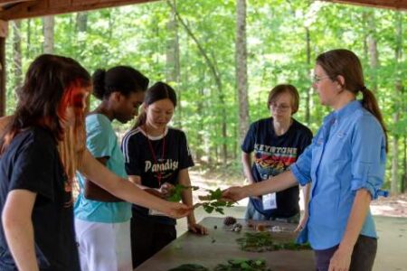 A group of four students stands around a table examining leaves with an instructor in an outdoor, wooded setting.
