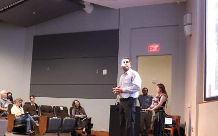 A man stands at a podium presenting to an audience in a lecture hall, with a woman nearby and a large projector screen displaying an image.