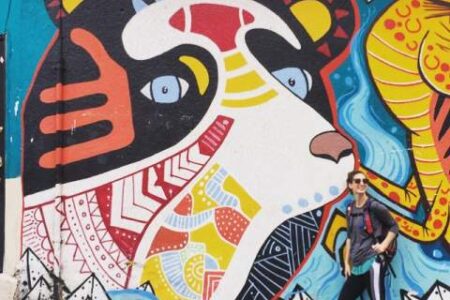 A person stands in front of a large, colorful mural of a bear painted on an outdoor wall.