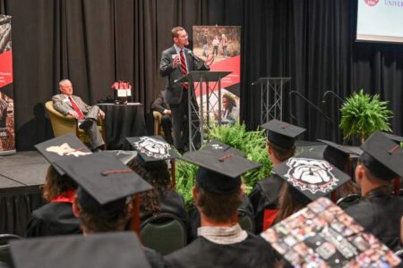 A speaker addresses graduates in caps and gowns at an indoor graduation ceremony, with an audience and banners visible in the background.