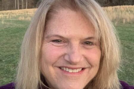 A woman with blonde hair smiles at the camera while standing outdoors in a grassy field with trees in the background.