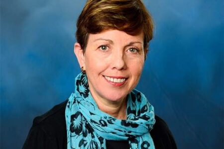 A woman with short brown hair wearing a black top and a blue floral scarf, smiling in front of a blue backdrop.