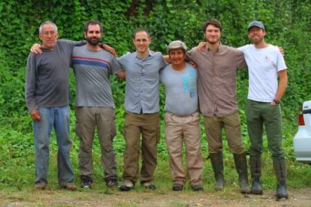 Six men stand outdoors in front of dense green foliage, posing side by side with arms around each other, some wearing boots and casual clothing.