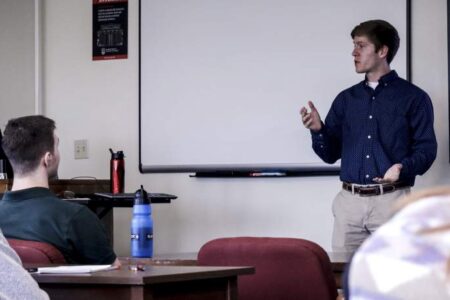 A man stands and speaks in front of a classroom, gesturing with one hand, while several students sit and listen. A whiteboard and presentation screen are visible behind him.