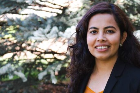 A woman with long curly hair, wearing a black blazer and orange top, smiles while standing outdoors in front of pine trees.