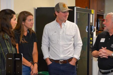 Four people stand indoors having a conversation; three are younger adults in casual clothes, and one is an older man in a black polo shirt with a name tag.