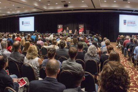 Large audience seated in a conference hall listens to a speaker on stage, with University of Georgia logos displayed on two projector screens at the front.