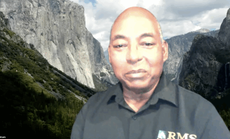 A man in a dark shirt with an RMS logo sits in front of a scenic mountain and forest background, likely a virtual backdrop.