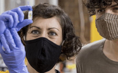 Two people wearing face masks look closely at a small vial held up by one person wearing purple gloves.