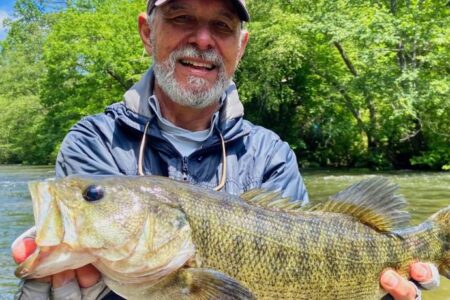 Man standing in a river, smiling and holding a large fish with both hands. Lush green trees are in the background.