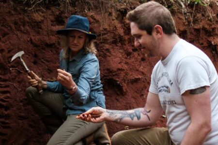 Two people examine soil samples at an outdoor excavation site; one holds a rock hammer and both are focused on items in their hands.