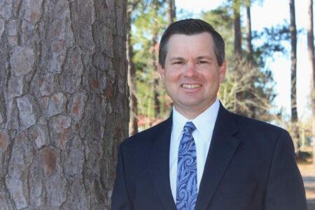 A man in a dark suit and patterned tie stands outdoors next to a large tree trunk, with trees and sunlight in the background.