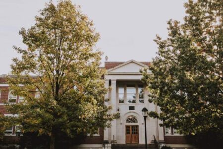 A brick and white stone building with large trees and stairs leading to the main entrance, photographed during daylight.