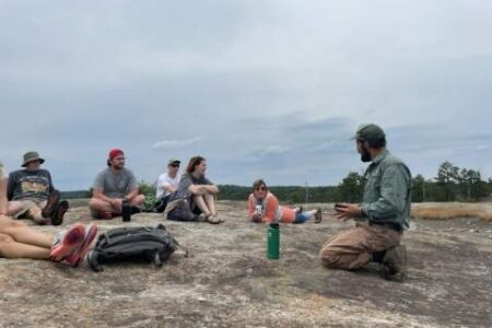 A group of people sit and listen to a person speaking while outdoors on a rocky surface under a cloudy sky.