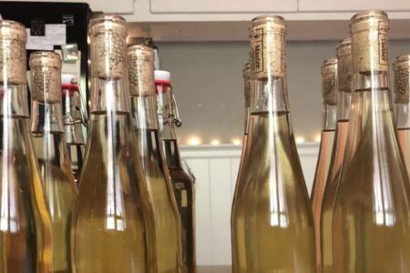 A row of clear glass bottles filled with a pale yellow liquid, sealed with corks, is displayed on a table indoors.