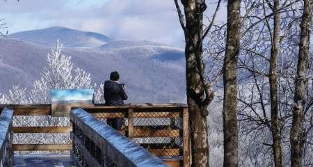 A person stands on a wooden overlook, taking a photo of snow-dusted mountains and frosty trees under a partly cloudy sky.